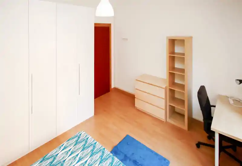 Bright, minimalist bedroom with a patterned bedcover, blue rug, wardrobe and a work desk — quiet and well-lit space ideal for a single occupant.