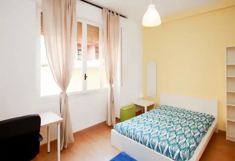 Bright, tidy bedroom with a double bed dressed in a patterned blue duvet, a bedside table and lamp, a small desk and chair by a large window letting in natural light.