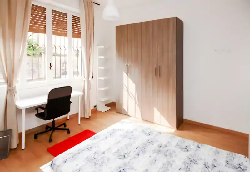 Sunny, minimalist bedroom with a double bed in the foreground, a white desk by the window and a large wooden wardrobe. Neutral curtains and warm wood flooring create a cozy, bright atmosphere ideal for relaxing or working.