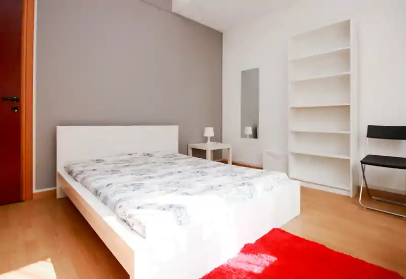 Bright, modern bedroom with a white low-profile bed, bedside table and lamp, a tall empty bookshelf and a red rug that adds a pop of color.