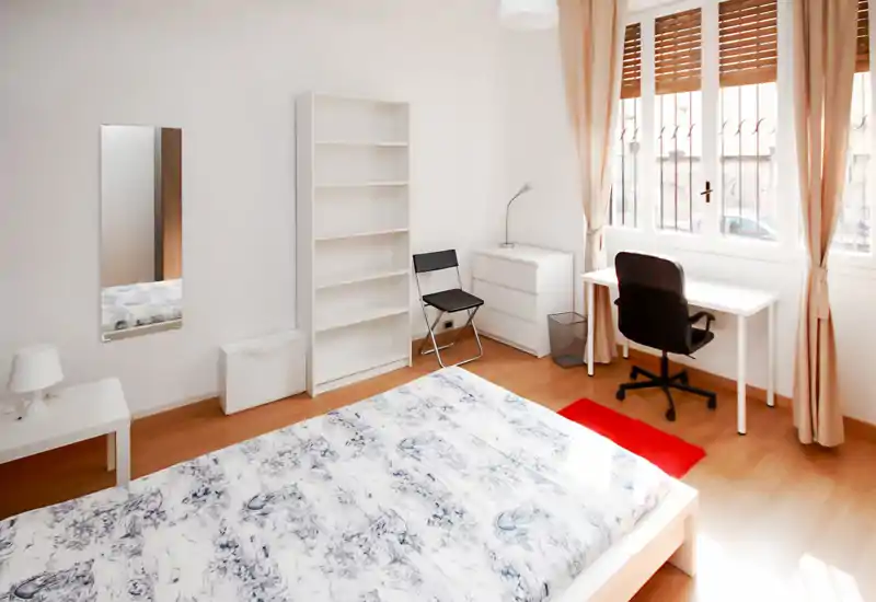 Bright, minimal bedroom with a large bed in the foreground, white shelving, desk by the window and warm wooden floors—ample natural light and simple, modern furnishings.