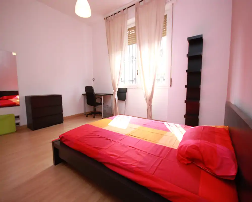 Bright, airy bedroom with a low-profile double bed dressed in red linens, a workspace by the window, dresser and modern shelving—ideal for a comfortable stay with natural light.