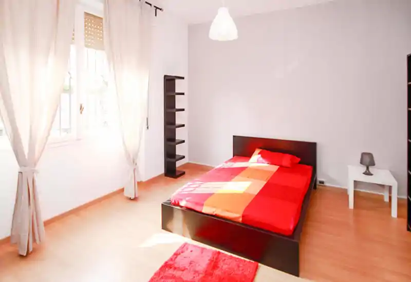 Bright, minimal bedroom with a double bed featuring red-orange linens, a bedside table with lamp, shelving and large windows that flood the room with natural light.