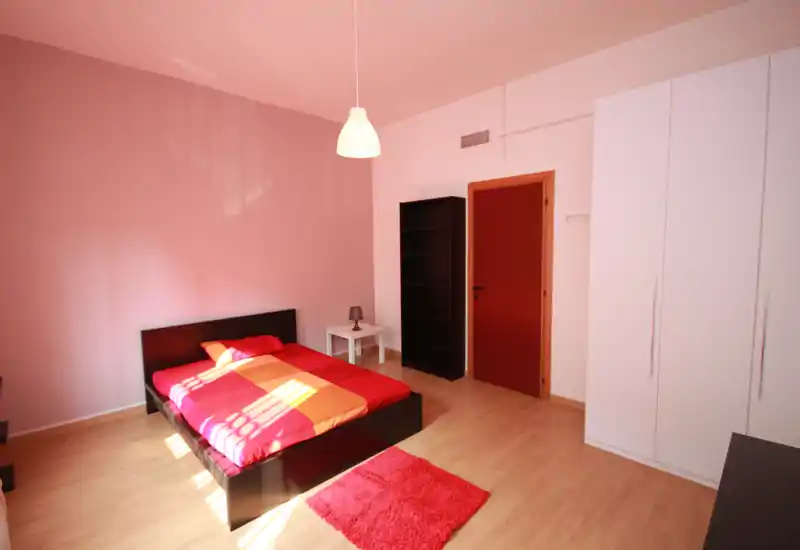 Spacious, bright bedroom with a double bed dressed in warm red and orange linens, a bedside table with lamp, wardrobe and shelving — ideal for comfortable stays.