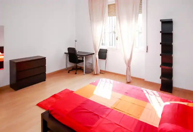 Bright, minimalist bedroom with a colorful double bed in the foreground, a desk by the window, dresser and vertical shelving — airy natural light and simple modern furniture.