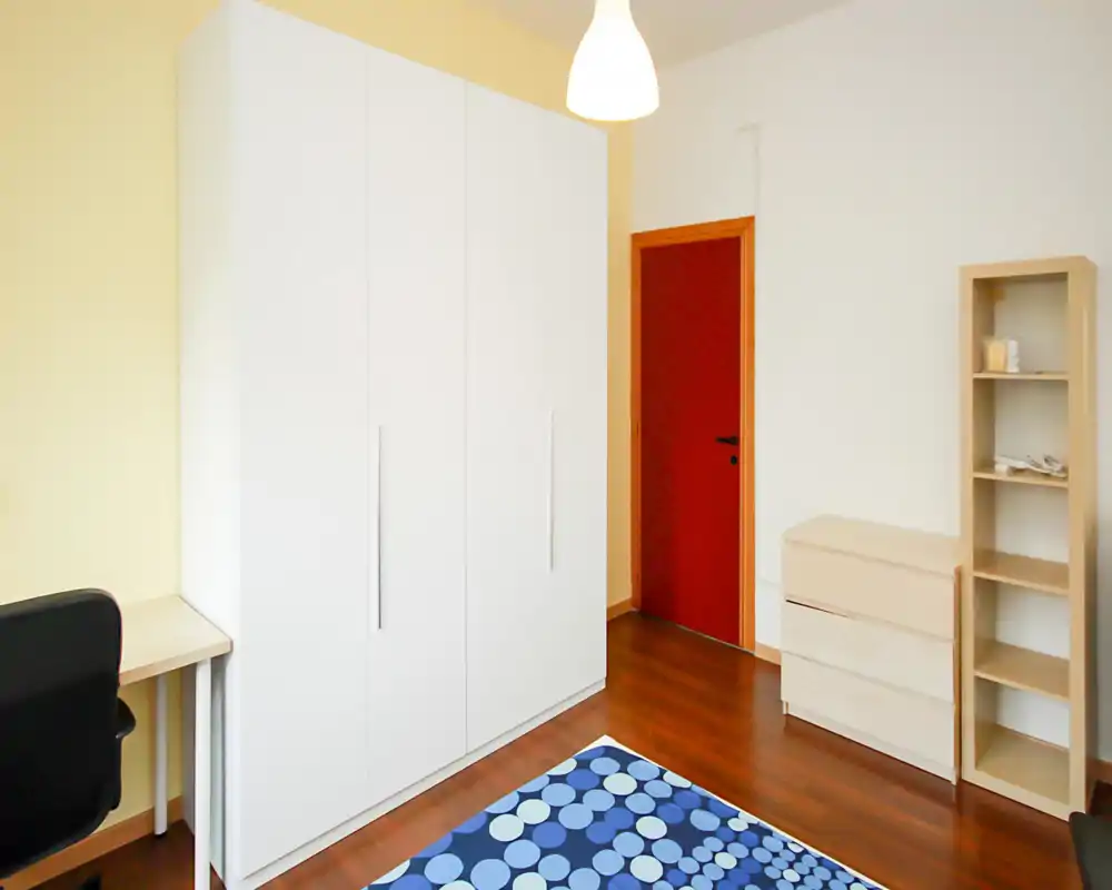 Angolo di camera da letto luminoso e minimale con un grande armadio bianco, cassetti e scaffale in legno chiaro e una coperta blu a motivo parzialmente visibile in primo piano.