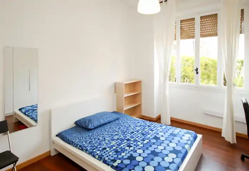 Camera da letto luminosa e minimalista con letto matrimoniale con copriletto blu a pois, ampia finestra con tende leggere e pavimento in legno che crea un’atmosfera ariosa e pulita.