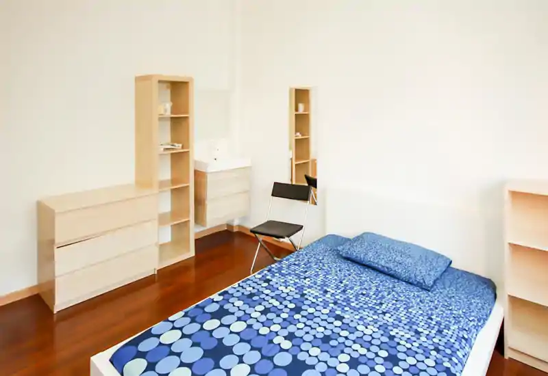 Camera da letto luminosa e minimal con letto matrimoniale dalla biancheria blu a motivo, mobili contenitori in legno chiaro e una sedia semplice — spazio moderno e ordinato ideale per il riposo.