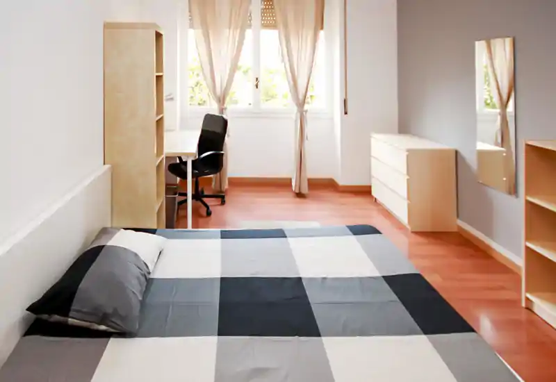 Bright, minimal bedroom with a large bed in the foreground, light wood furniture, a desk by the window and soft natural light creating a calm atmosphere.