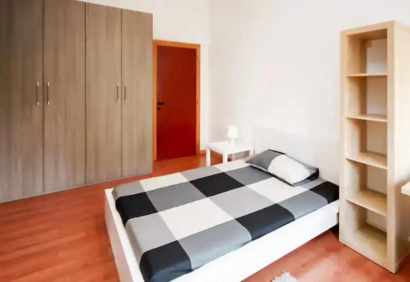 Clean, minimal bedroom with a double bed dressed in a gray-and-white checkered duvet, a bedside table with lamp, large wardrobe and open shelving — bright and simply furnished.