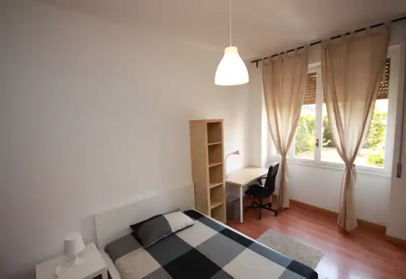 Bright, tidy bedroom with a double bed, bedside table, shelving and a desk by a large window letting in natural light.