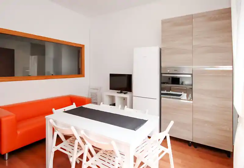 Bright, modern living area with a compact dining table, white chairs and a vivid orange sofa next to built-in kitchen cabinets and a fridge — ideal for a small apartment.