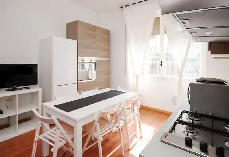 Cocina moderna y luminosa con mesa de comedor para seis, horno empotrado y frigorífico; suelo de madera y muebles blancos que crean un ambiente limpio y aireado.