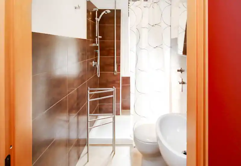 Baño moderno y luminoso con zona de ducha, lavabo y WC blancos, cortina de ducha con estampado y toallero térmico cromado; azulejos limpios y grifería contemporánea.