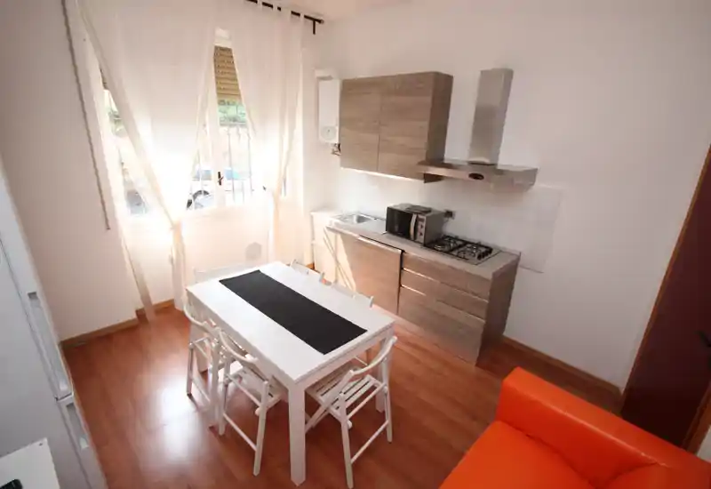 Cocina abierta, compacta y luminosa con mesa de comedor, muebles de madera, encimera a gas y una gran ventana que aporta luz natural — ideal para un piso pequeño.