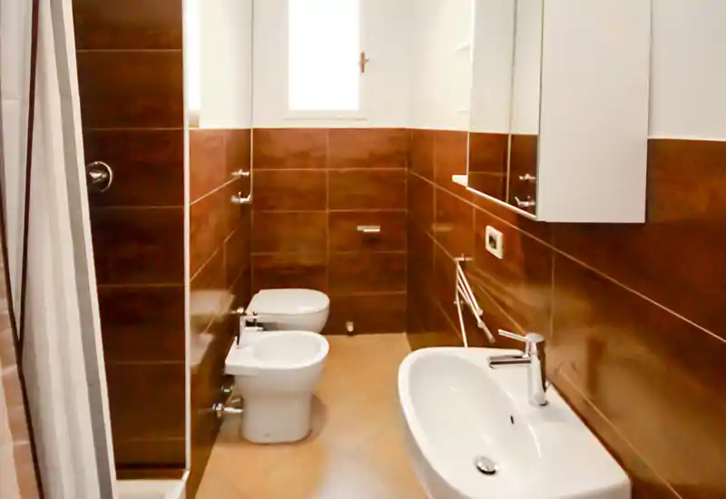 Baño moderno y luminoso con azulejos en la pared, bidé y WC, ducha separada con cortina y un lavabo blanco con armario con espejo.