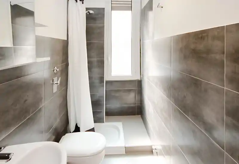 Baño moderno y compacto con azulejos grises, plato de ducha con cortina, inodoro y lavabo pequeño; luz natural intensa por una ventana estrecha.