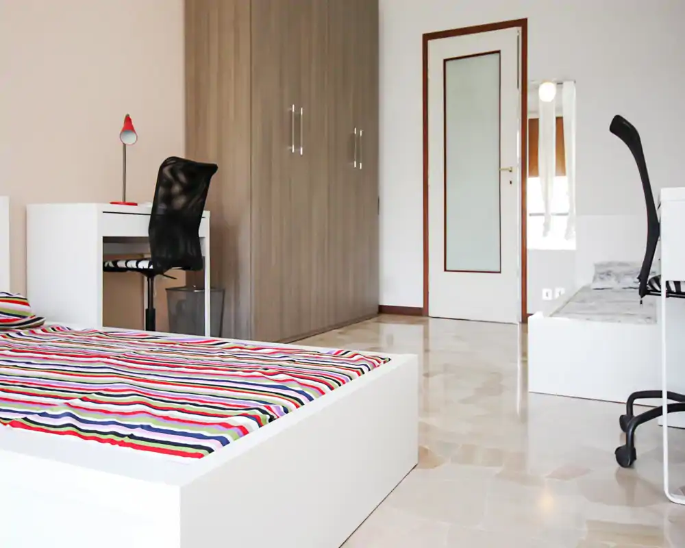 Dormitorio moderno y luminoso con una cama baja blanca y ropa de cama a rayas coloridas, escritorio y silla junto a un gran armario — estilo minimalista y mucha luz natural.