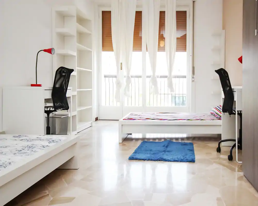 Dormitorio luminoso y minimalista con dos camas bajas blancas, gran ventana-balcon que aporta luz natural y escritorios con estanterías — espacio fresco y ventilado, ideal para estudiantes o alquiler compartido.
