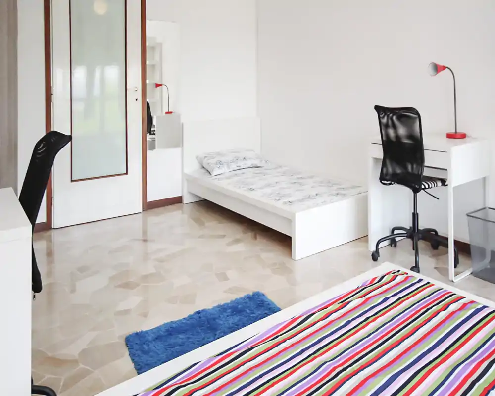 Dormitorio luminoso y minimalista con una cama blanca, un escritorio con silla y una cama de rayas coloridas en primer plano. La habitación es amplia, ordenada y bien iluminada, ideal para anuncios que resaltan la simplicidad y un área de estudio.