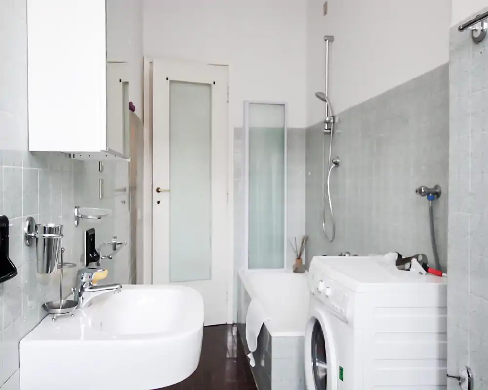 Baño luminoso y limpio con lavabo, bañera con ducha y lavadora de carga frontal; azulejos gris claro y sanitarios blancos para un aspecto minimalista.