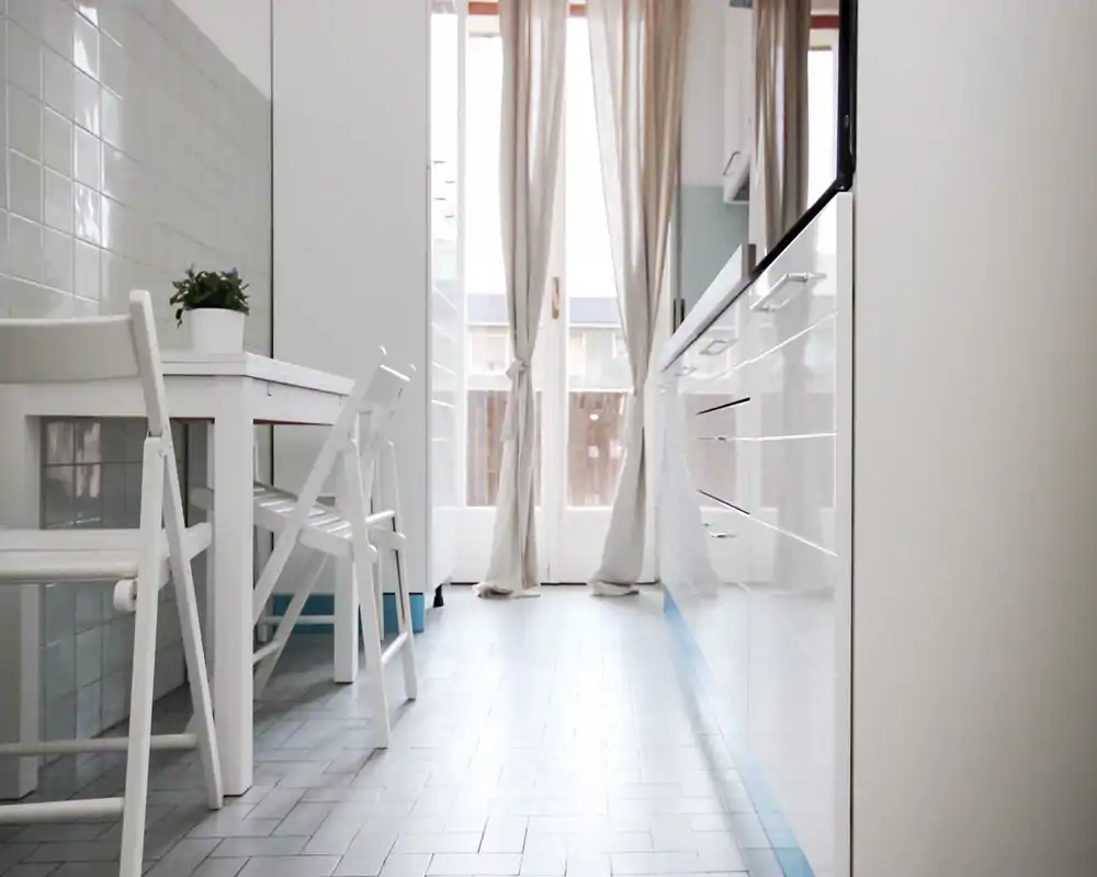 Cocina moderna y luminosa con armarios blancos brillantes, una mesa blanca pequeña con sillas plegables y luz natural que entra por la puerta acristalada con cortinas.
