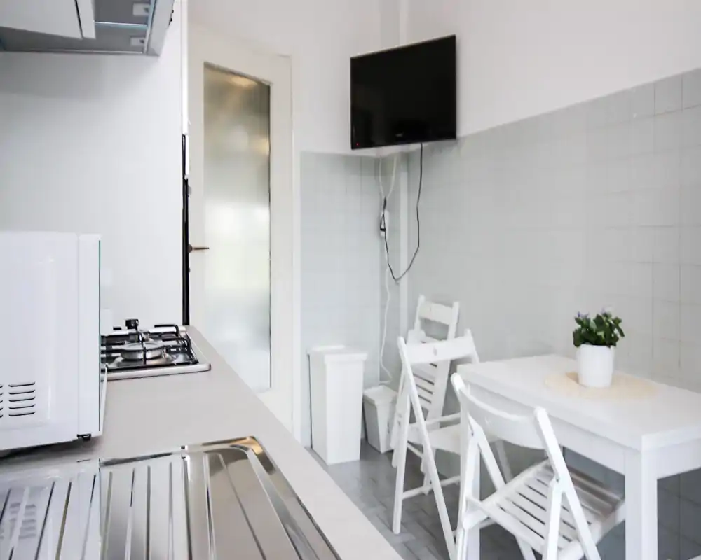 Cocina compacta y luminosa con placa de gas, microondas y pequeña mesa de comedor. Estilo limpio y minimalista con muebles blancos y una planta en maceta que añade un toque verde.