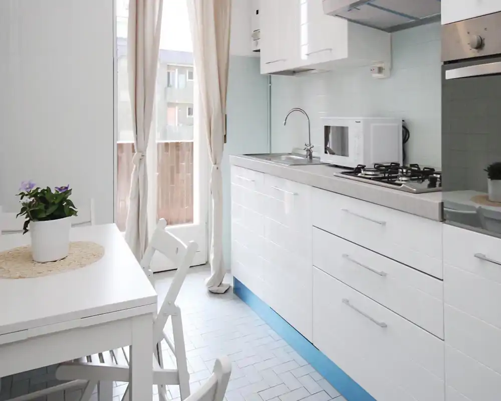 Cocina moderna y luminosa con muebles blancos, vitrocerámica a gas, microondas y una pequeña mesa junto a una puerta soleada; detalles frescos como una planta en maceta y cortinas neutras.
