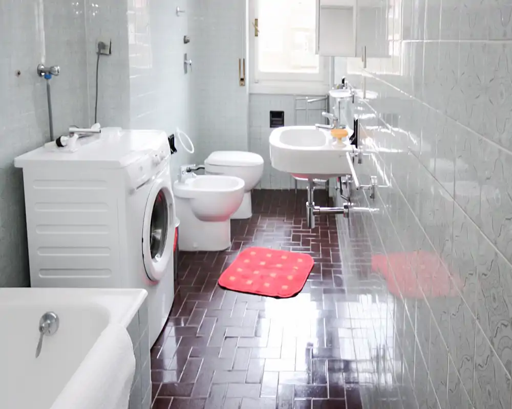 Baño luminoso con bañera, lavadora, bidé, inodoro y lavabo de pared; azulejos brillantes y una pequeña alfombra de baño roja aportan contraste.