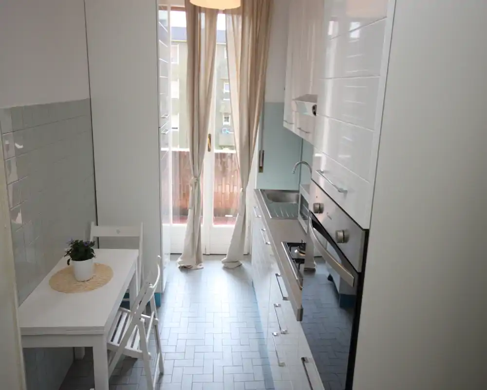 Cocina estrecha y luminosa tipo galera con muebles blancos, electrodomésticos integrados y una pequeña mesa junto a la puerta ventana; la luz natural aporta sensación de limpieza y frescura.
