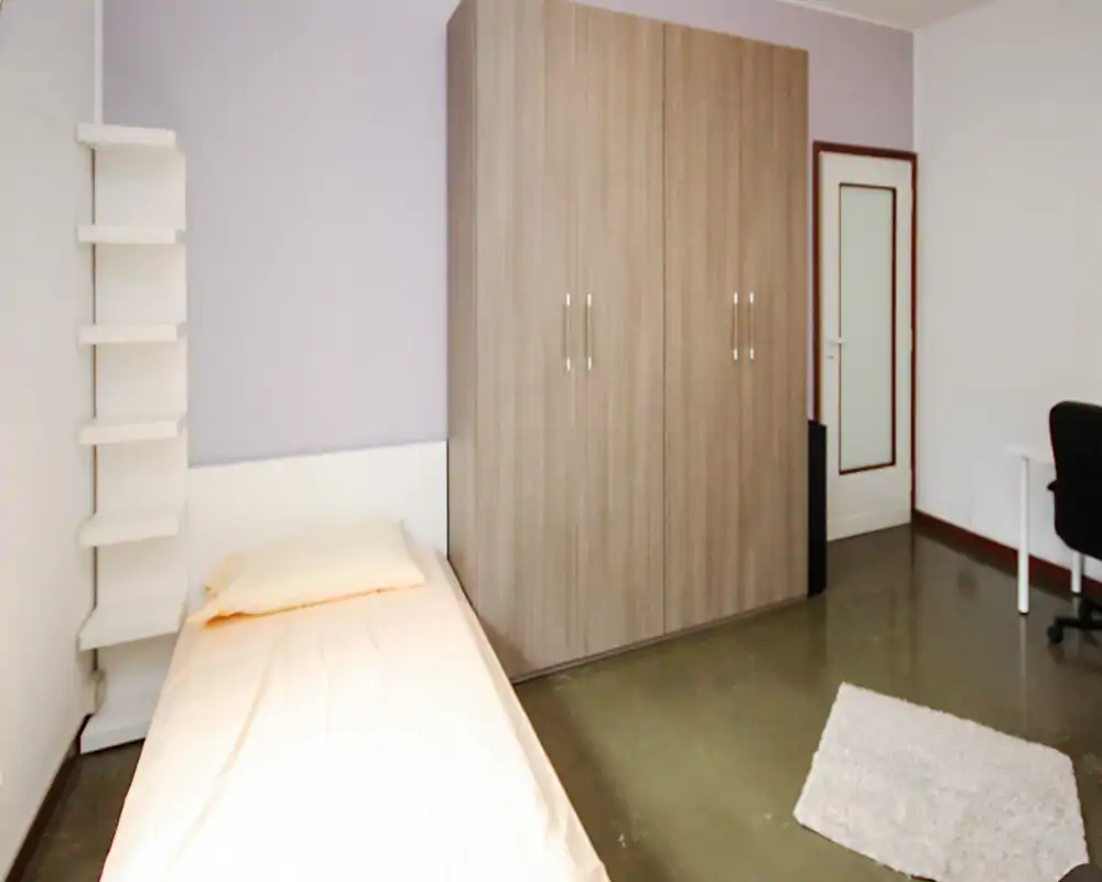 Camera singola semplice e ordinata con letto rifatto, ampio armadio e piccolo angolo scrivania; toni neutri e arredo essenziale per uno spazio funzionale e tranquillo.