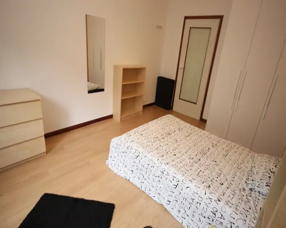 Camera da letto minimal e ben illuminata con letto matrimoniale con copriletto stampato, armadio, comò e piccolo scaffale — arredamento neutro ideale per un annuncio di affitto.