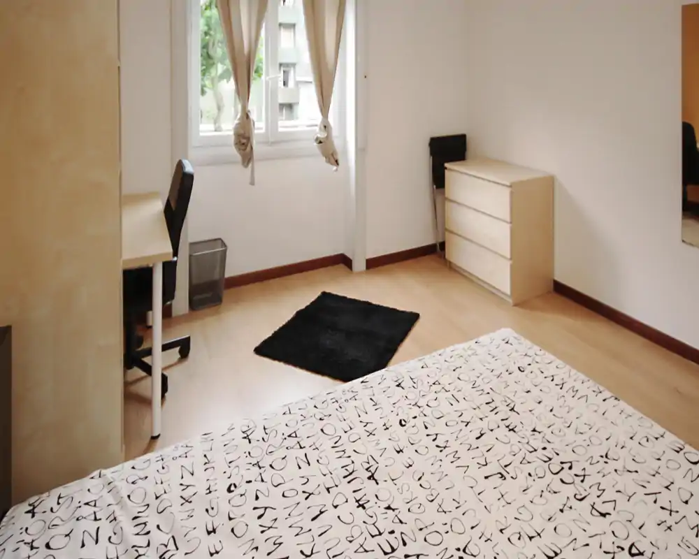 Camera da letto luminosa e minimal con copriletto stampato in primo piano, scrivania e sedia vicino alla finestra e comò in legno chiaro. Ambiente arioso e arredamento moderno, adatto per un annuncio di affitto.