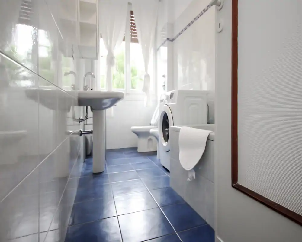 Bagno luminoso e pulito con pavimento piastrellato blu, lavabo a colonna, WC e lavatrice accanto alla vasca; luce naturale dalla finestra con tende.