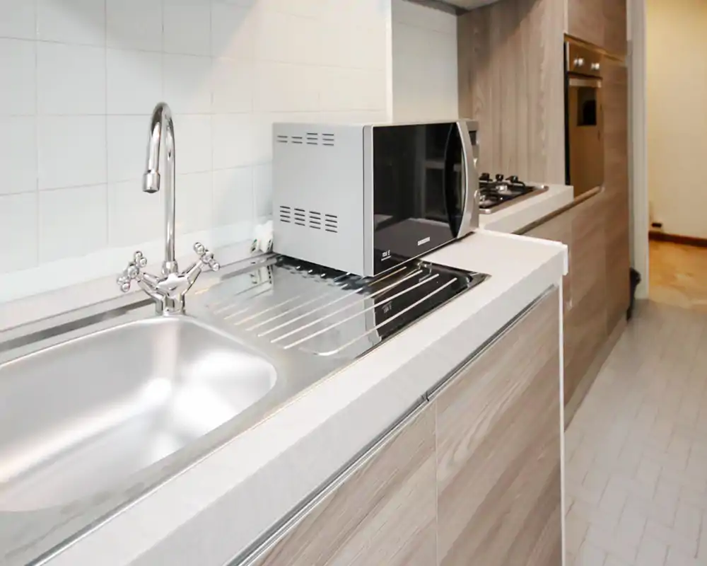 Cucina moderna compatta con lavello in acciaio inox, piano cottura integrato e microonde su piano in legno chiaro — luminosa, pulita e funzionale.
