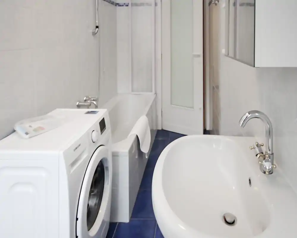 Bagno moderno e luminoso con vasca, lavabo con miscelatore cromato e lavatrice a carica frontale; piastrelle bianche e pavimento blu donano un aspetto pulito.