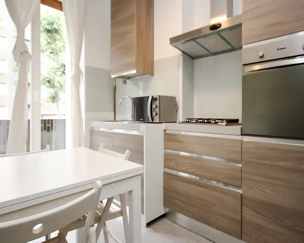 Cucina moderna e luminosa con mobili effetto legno, piano cottura a gas, forno incassato e tavolo bianco vicino a una grande finestra che illumina l'ambiente.
