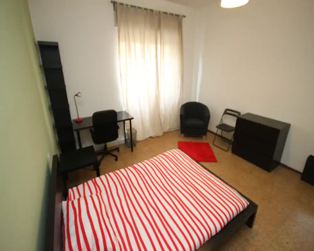 Dormitorio luminoso y sencillo con cama doble con ropa de cama a rayas rojas y blancas, escritorio junto a la ventana, sillón y cómoda.