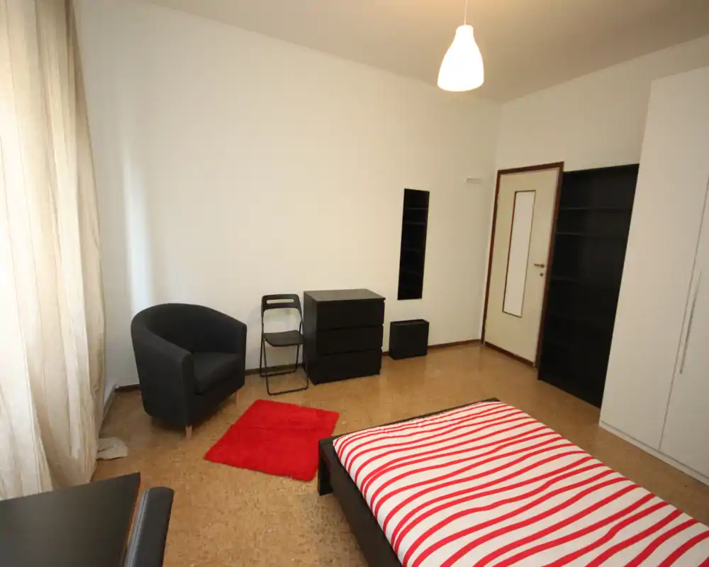 Dormitorio luminoso y amueblado de forma sencilla con cama doble con ropa de cama a rayas rojas y blancas, un sillón, estanterías y luz natural de una gran ventana.