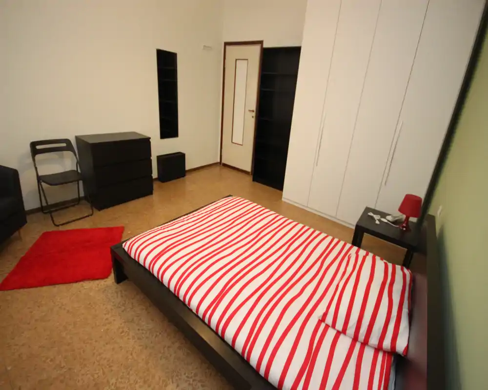 Dormitorio luminoso y bien presentado con una cama doble grande con ropa de cama a rayas rojas y blancas, mesita con lámpara, armario empotrado y mobiliario moderno sencillo.