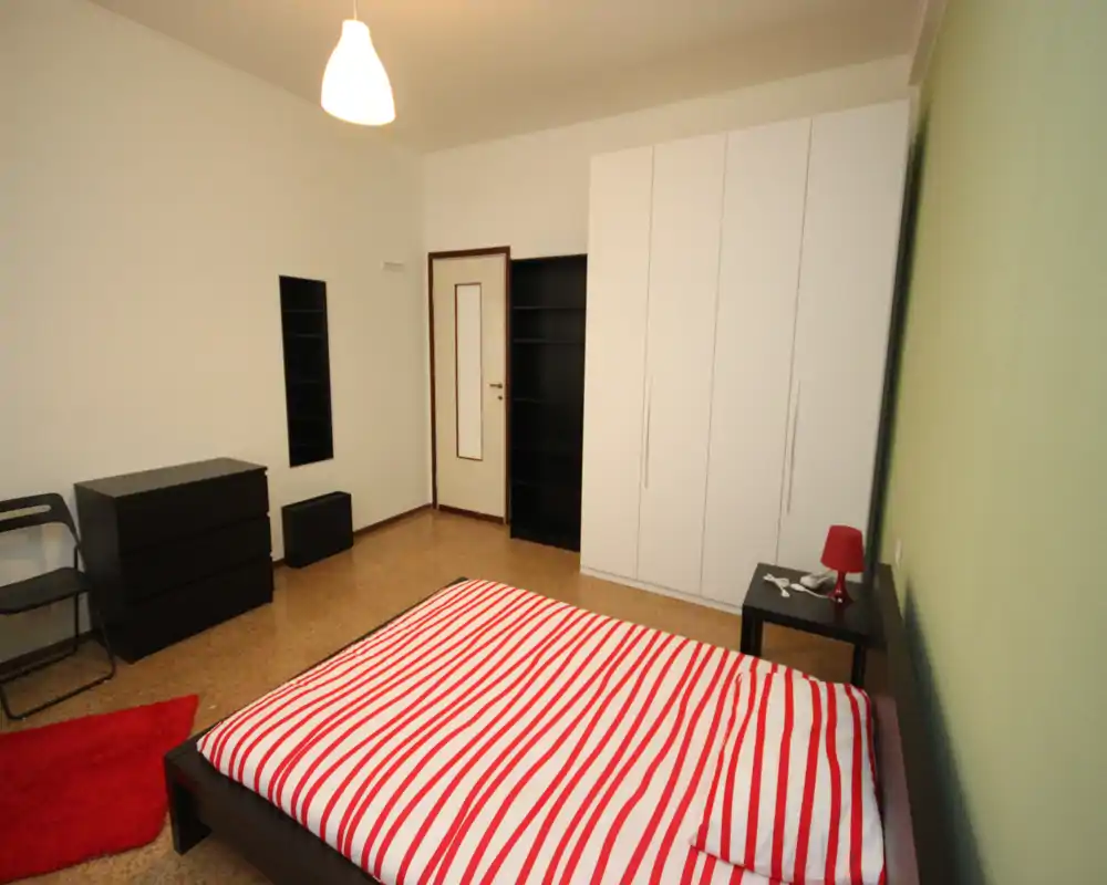 Dormitorio luminoso y sencillo con cama doble con funda de rayas rojas y blancas, amplio armario blanco, mesita con lámpara y decoración mínima — perfecto para un anuncio limpio y moderno.