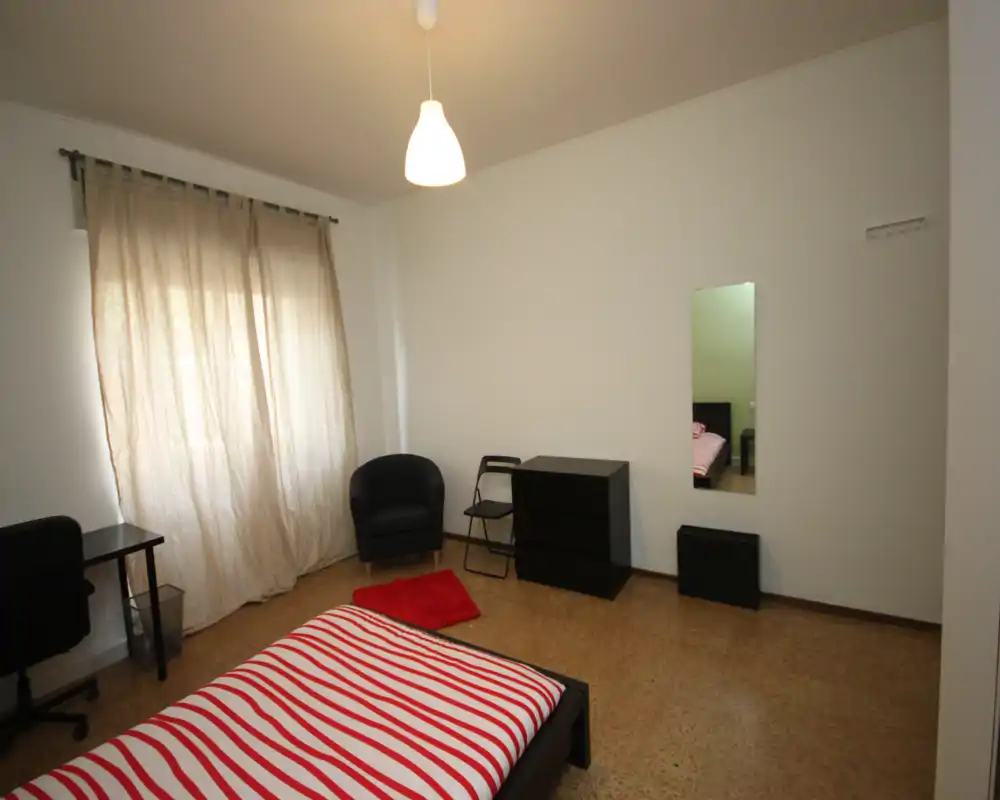 Dormitorio luminoso y amueblado de forma sencilla con cama doble con ropa de cama a rayas, mesa, sillón y cómoda. Gran ventana con cortina que aporta luz natural suave y paredes neutras.