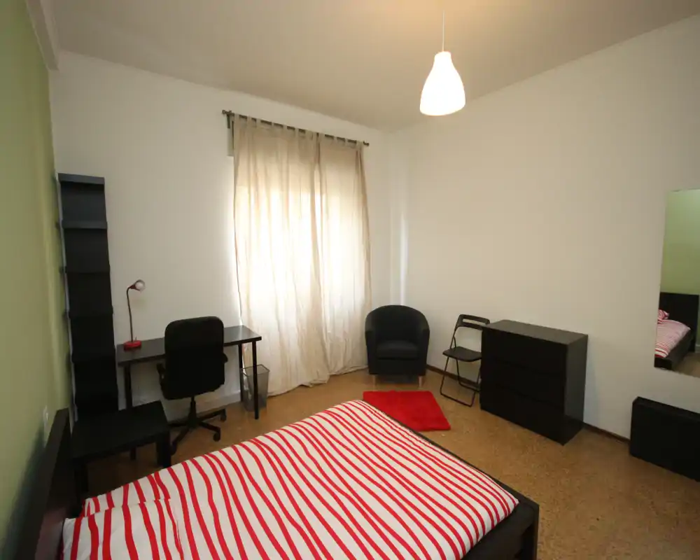 Dormitorio amplio y de mobiliario sencillo con cama doble con ropa de cama roja y blanca a rayas, escritorio junto a la ventana, butaca y cómoda — luz natural y decoración minimalista.