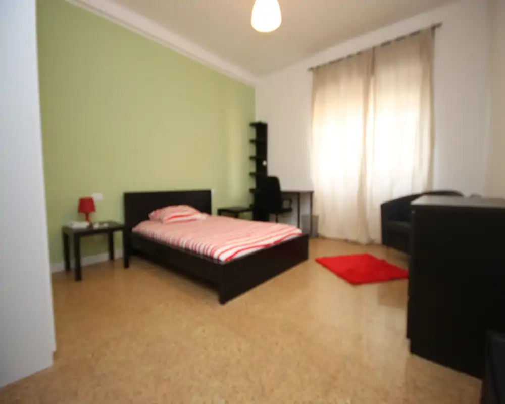 Dormitorio luminoso y de estilo sencillo con cama doble, mesita y escritorio. Las cortinas claras difunden la luz natural y la pared verde aporta un toque moderno y fresco.