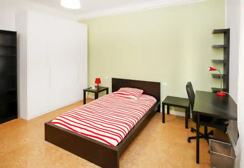 Dormitorio individual luminoso y moderno con estructura de cama oscura y ropa de cama a rayas rojas y blancas, mesita y zona de escritorio — ideal para estudiante o persona sola.