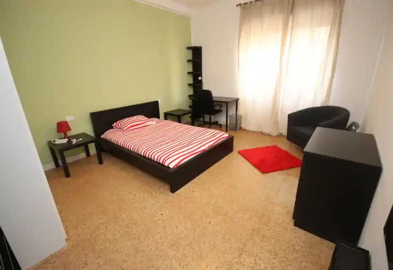 Dormitorio luminoso y ordenado con cama doble, escritorio y zona de estar; pared verde y ropa de cama a rayas que aportan un aire moderno y fresco.