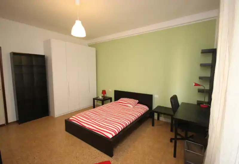 Dormitorio individual luminoso y ordenado con cama de madera oscura y ropa de cama a rayas rojas, mesita y escritorio; pared verde y amplio armario.