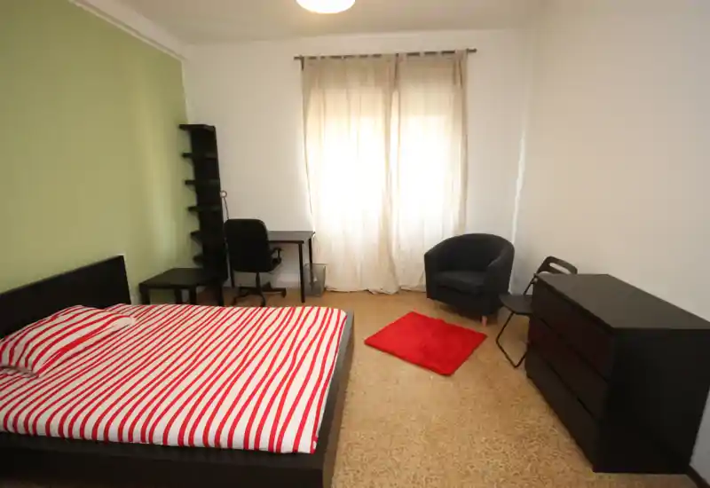 Dormitorio luminoso y bien distribuido con cama doble, escritorio, butaca y cómoda; los detalles rojos del cojín y la alfombra añaden calidez.