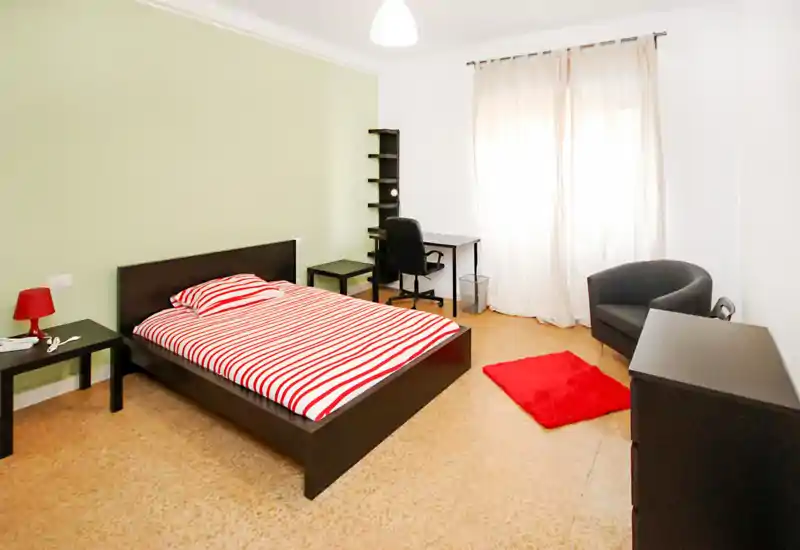 Dormitorio luminoso y moderno con cama doble, ropa de cama a rayas, mesita y una zona de trabajo junto a la ventana. Dispone de un sillón cómodo y mobiliario sencillo y coordinado, ideal para estudiantes o viajeros.