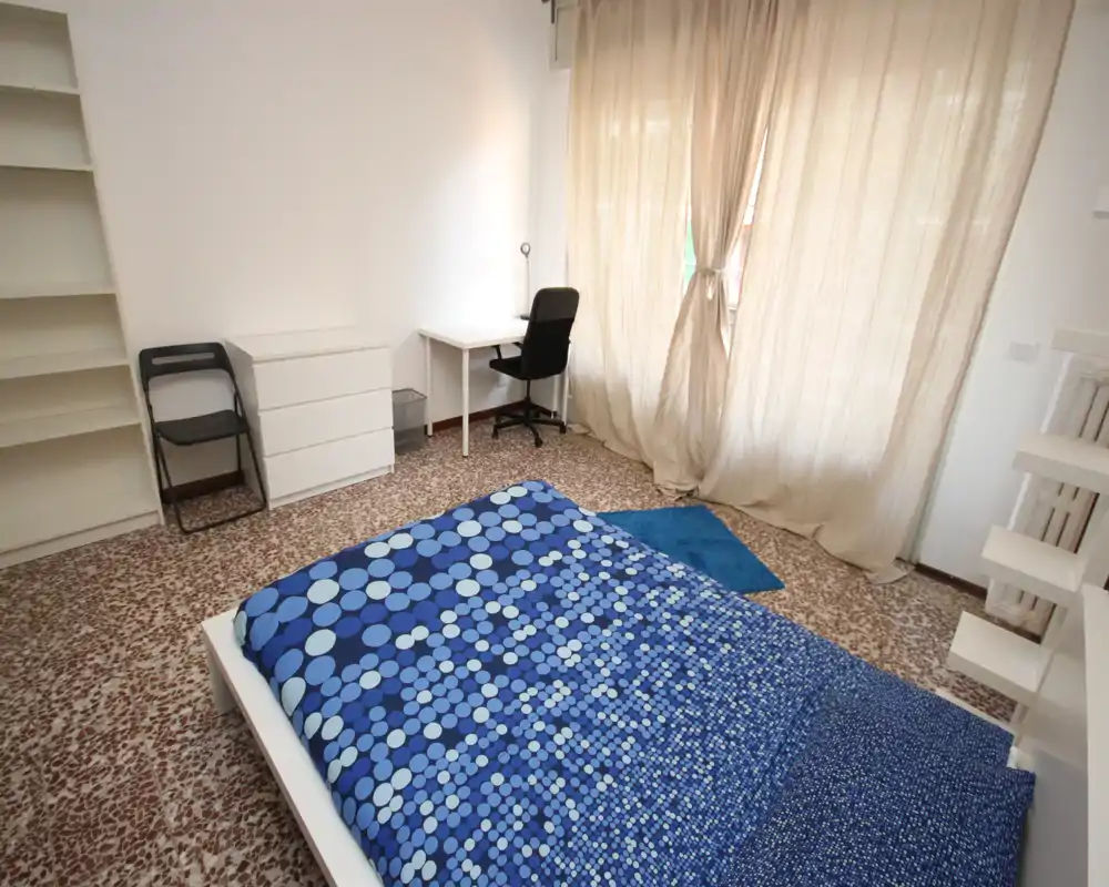 Bright, minimal bedroom with a double bed dressed in blue patterned bedding, a small desk and chair by a window with light curtains, and shelving for storage.